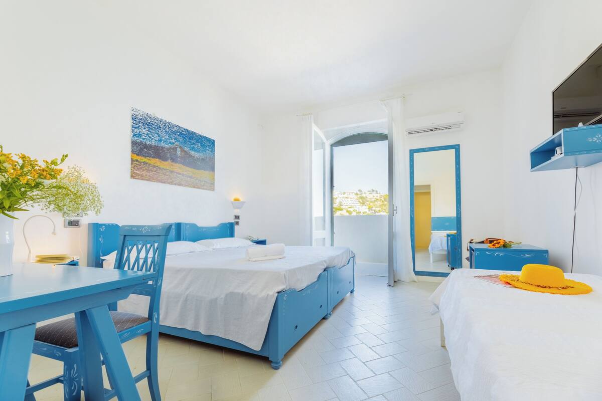 A room with a bed, a desk, and a chair. There is a painting on the wall and a window with curtains.
