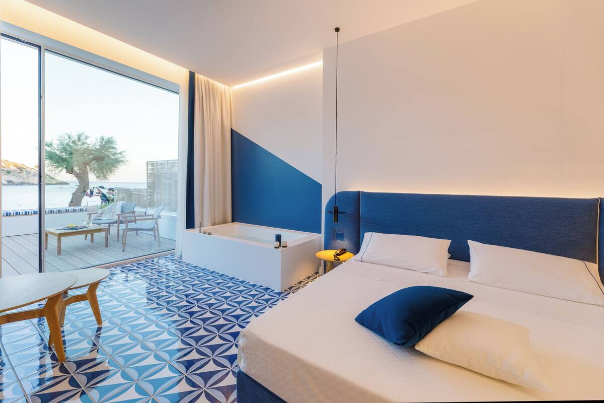 A modern hotel room with a large bed, a separate bathroom, a balcony with a view of the sea, and a dining area with a table and chairs.