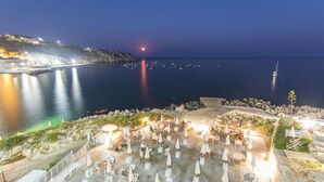 A coastal outdoor dining area with tables and chairs, overlooking a calm sea and a rocky shoreline.