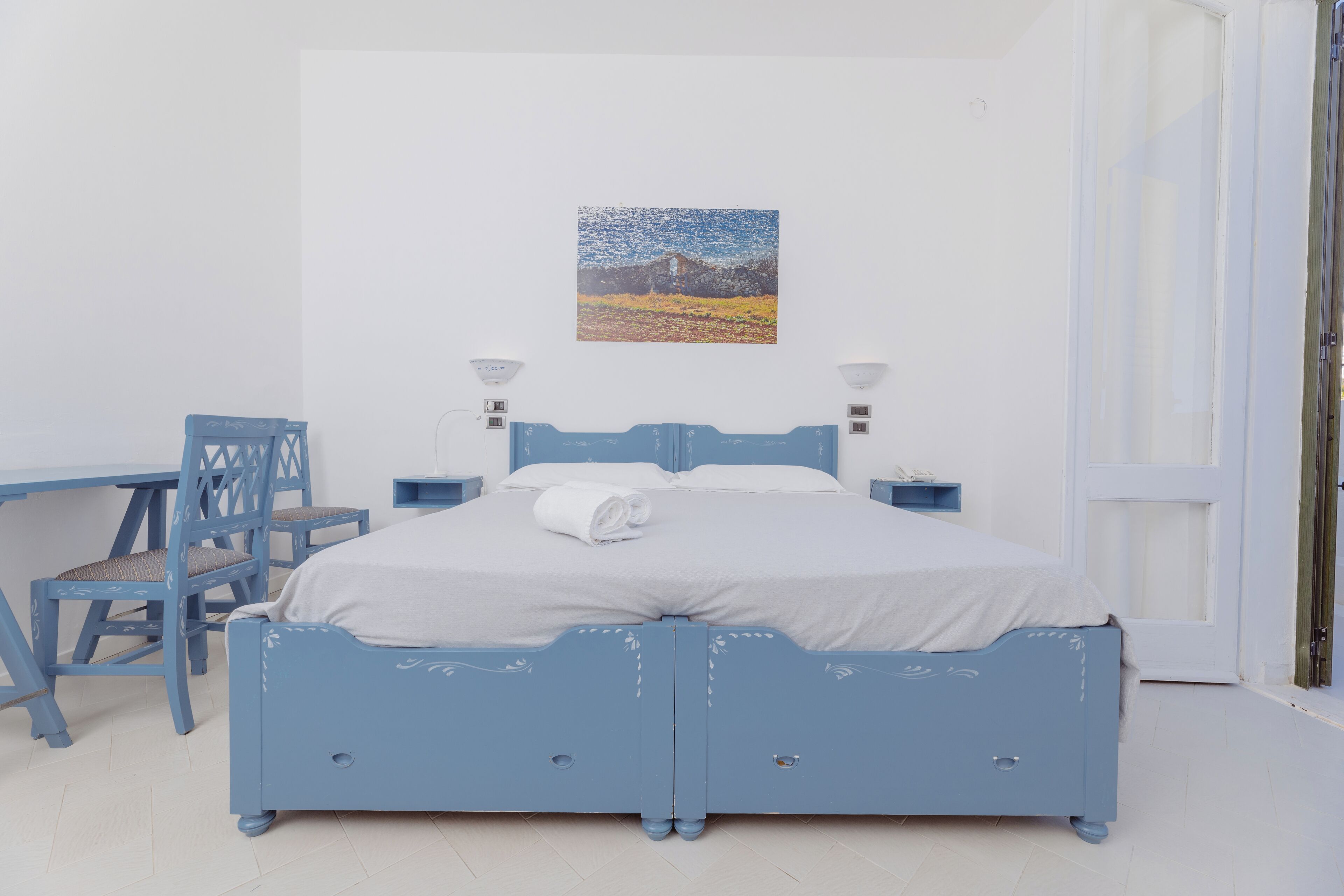 Un dormitorio con cama, escritorio y silla. Hay un cuadro en la pared y una puerta a la derecha.