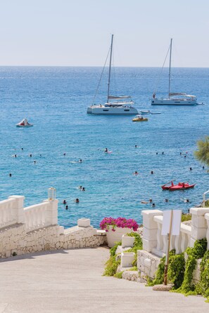 A coastal scene with a stone pathway leading to the sea, where several boats are anchored and people are swimming.