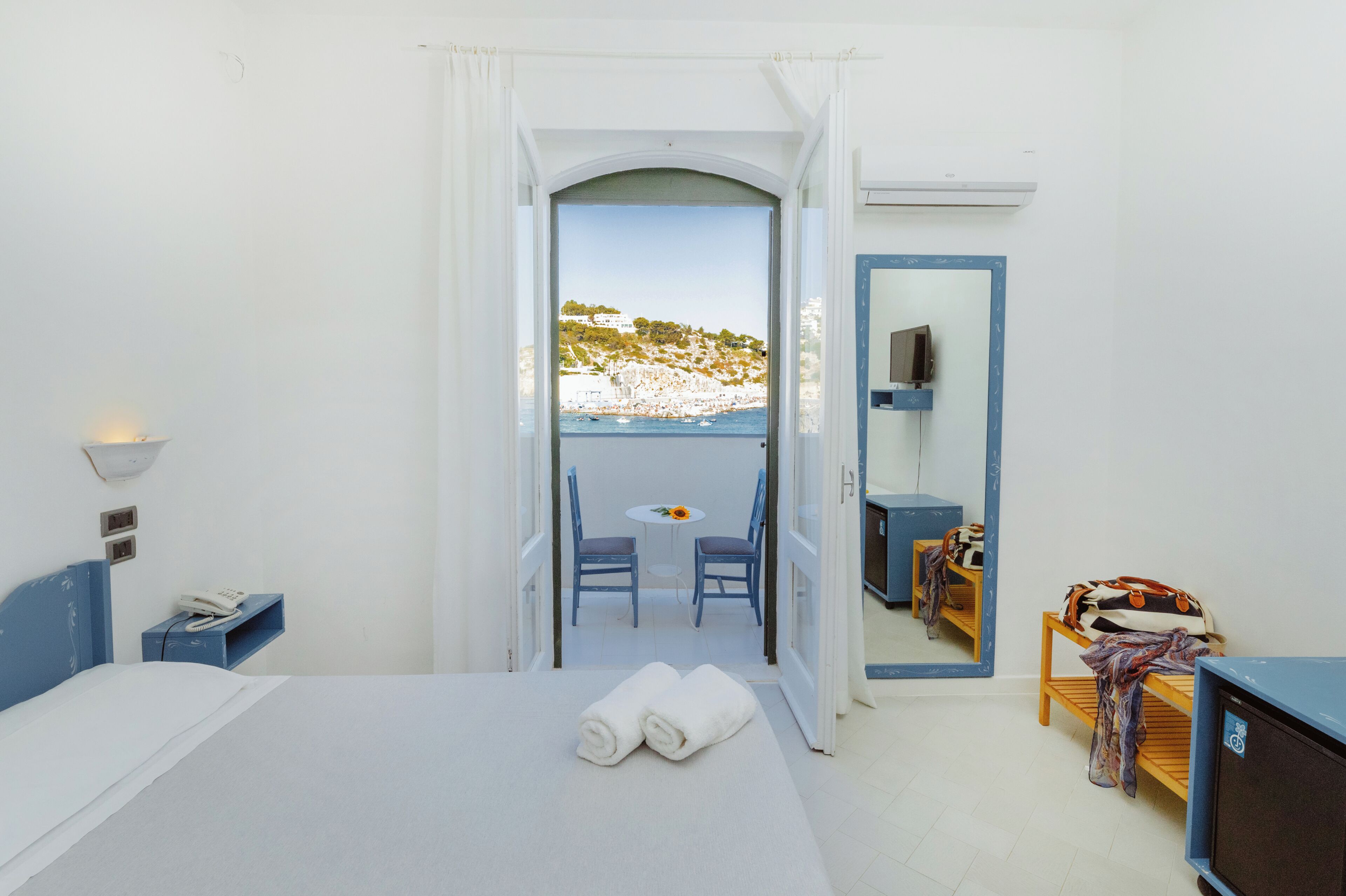 A room with a bed, a small table with chairs, a TV, and a balcony with a view of the sea and buildings.
