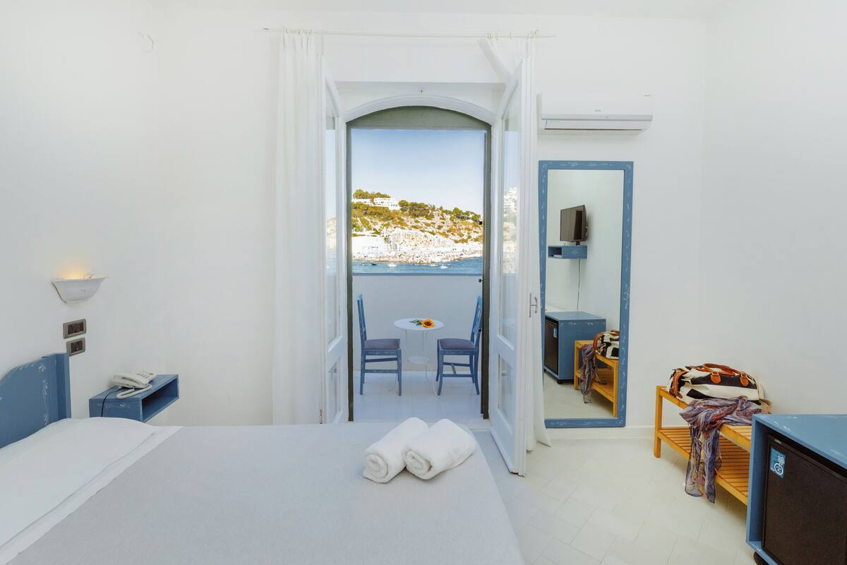 A hotel room with a bed, a small table with chairs, a TV, and a balcony with a view of the sea and a hill.
