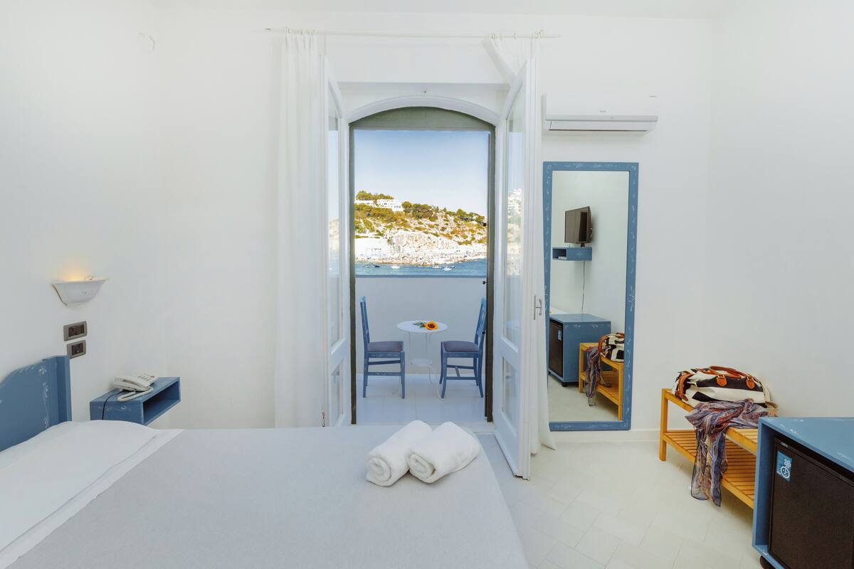 A hotel room with a bed, a small table with chairs, a TV, and a balcony with a view of the sea and a hill.