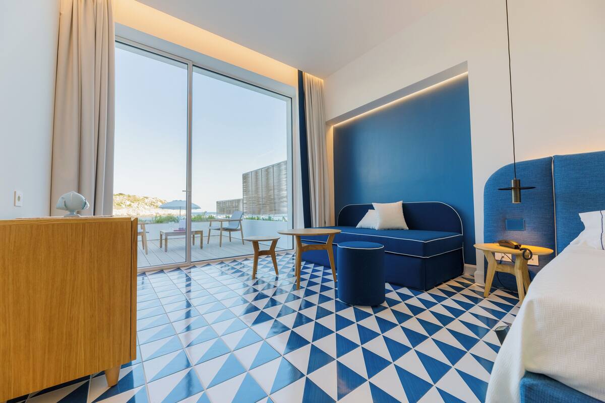 A hotel room with a large window, a blue sofa, a wooden desk, and a patterned floor.