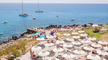 A beachside resort with multiple sun umbrellas, lounge chairs, and a rocky shoreline.