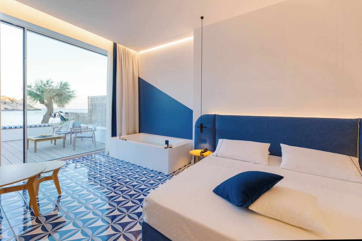A modern hotel room with a large bed, a separate bathroom, a balcony with a view of the sea, and a dining area with a table and chairs.