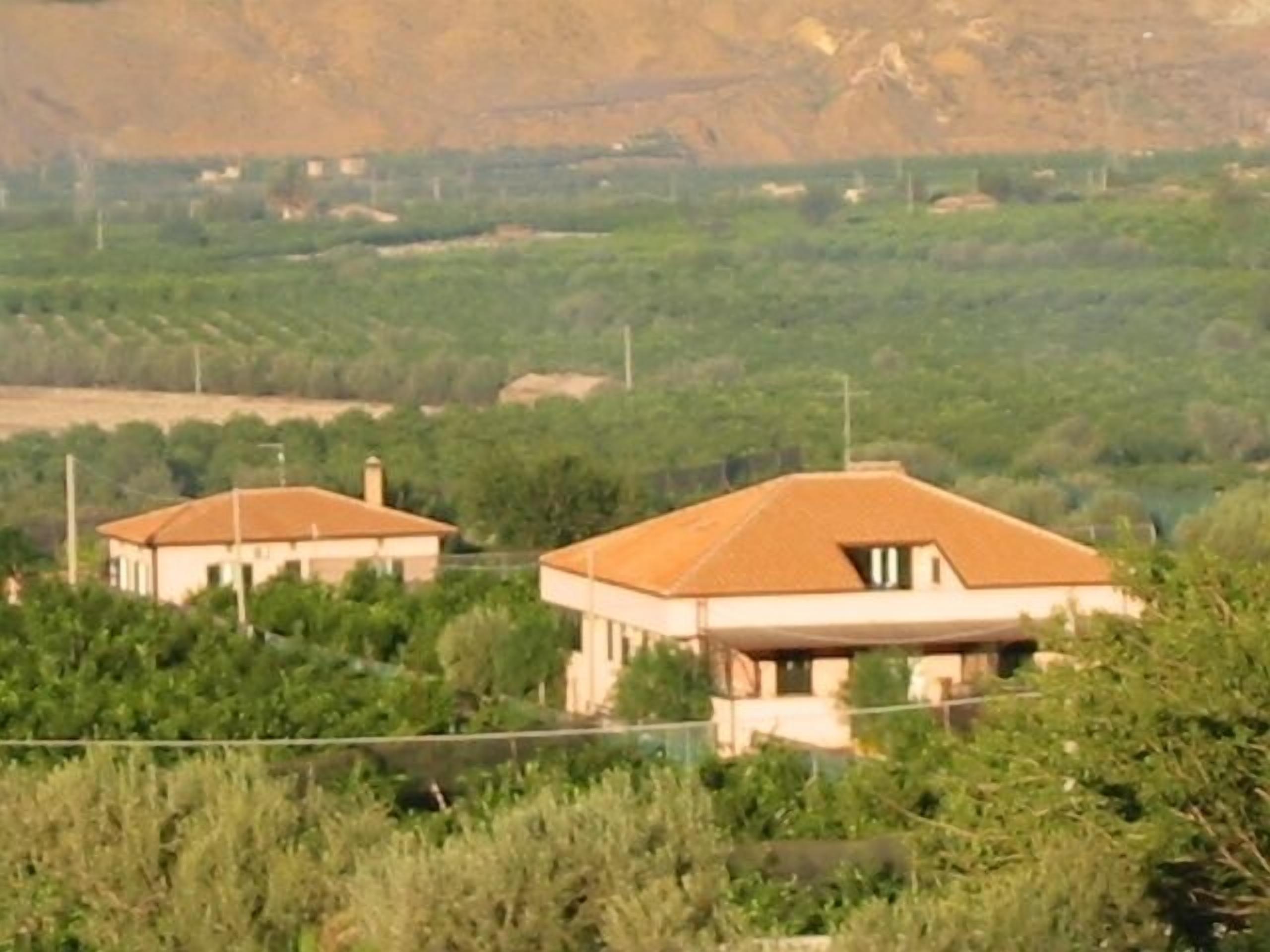 A rural setting with two houses surrounded by greenery and a hilly landscape in the background.