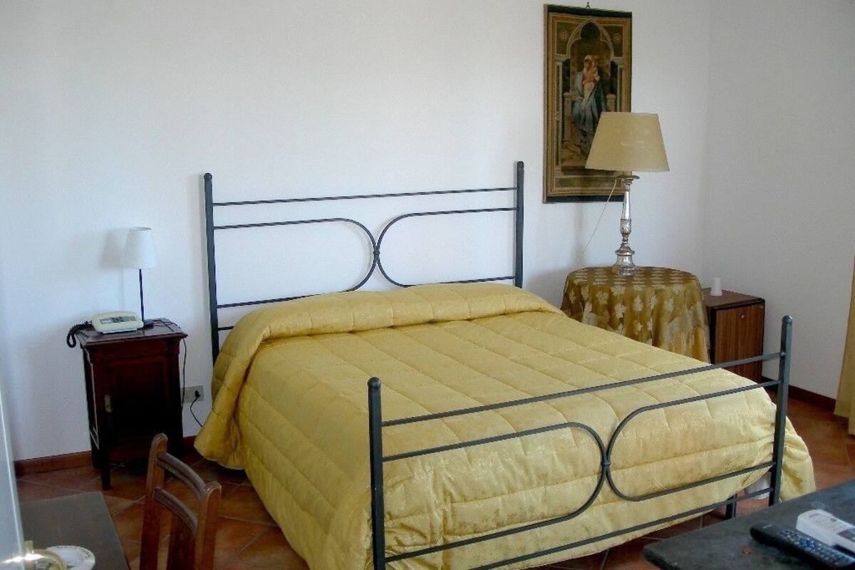A single-bed room with a metal frame, a yellow bedspread, a bedside table with a lamp, a small wooden nightstand, and a framed picture on the wall.