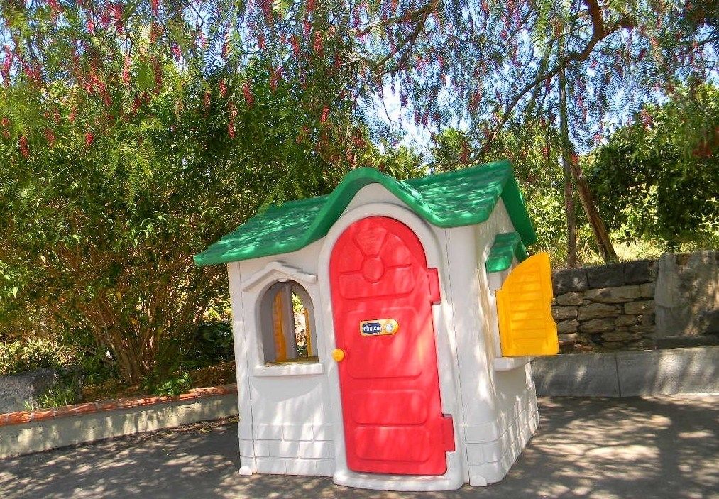 A small, white playhouse with a red door and green roof, surrounded by trees.