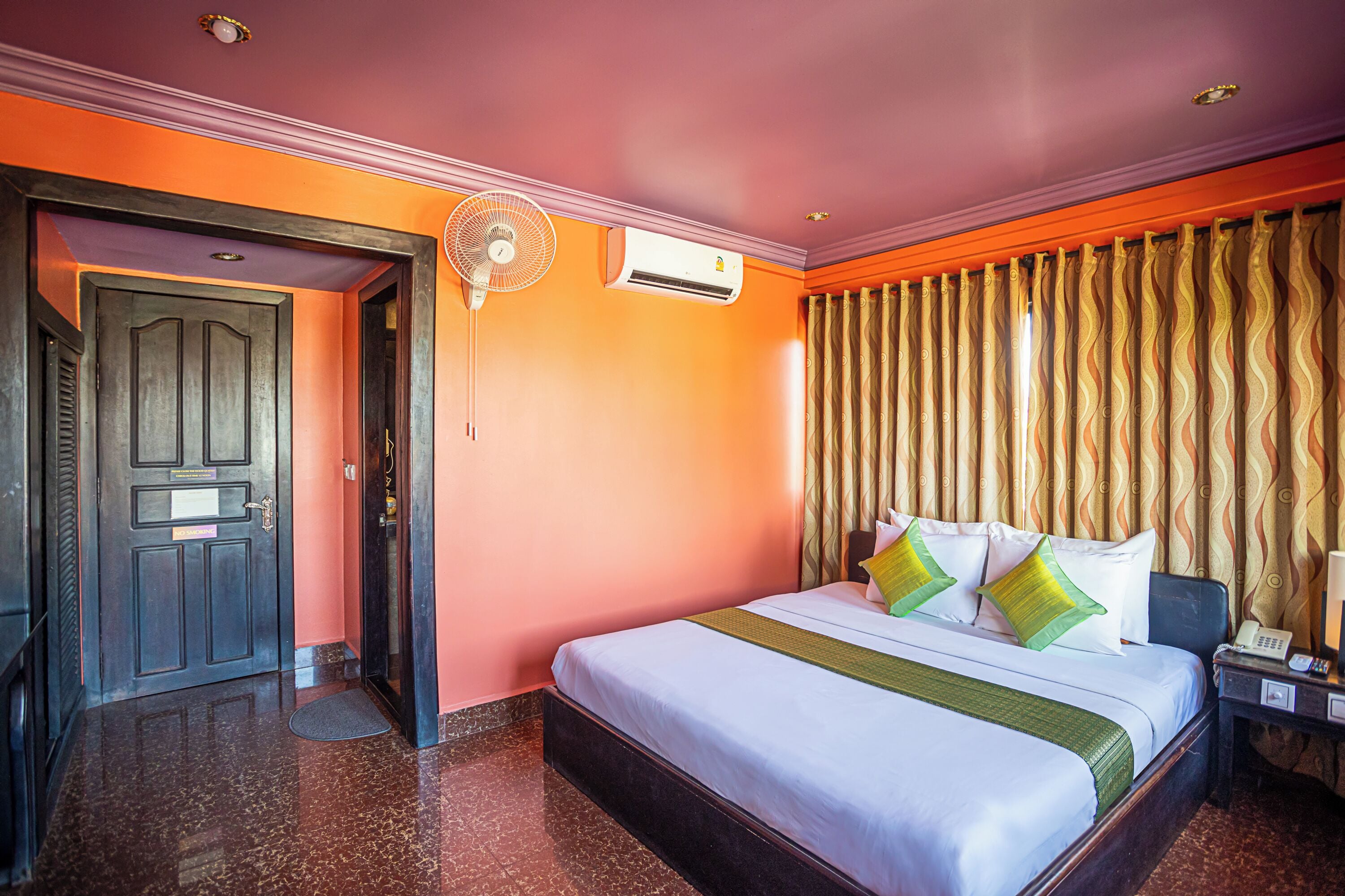 A hotel room with a bed, an air conditioning unit, a fan, a nightstand, and a door.