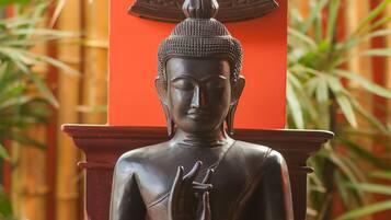 Una estatua sentada de Buda en posición meditativa, colocada sobre una plataforma de madera con un fondo rojo y plantas de bambú al fondo.