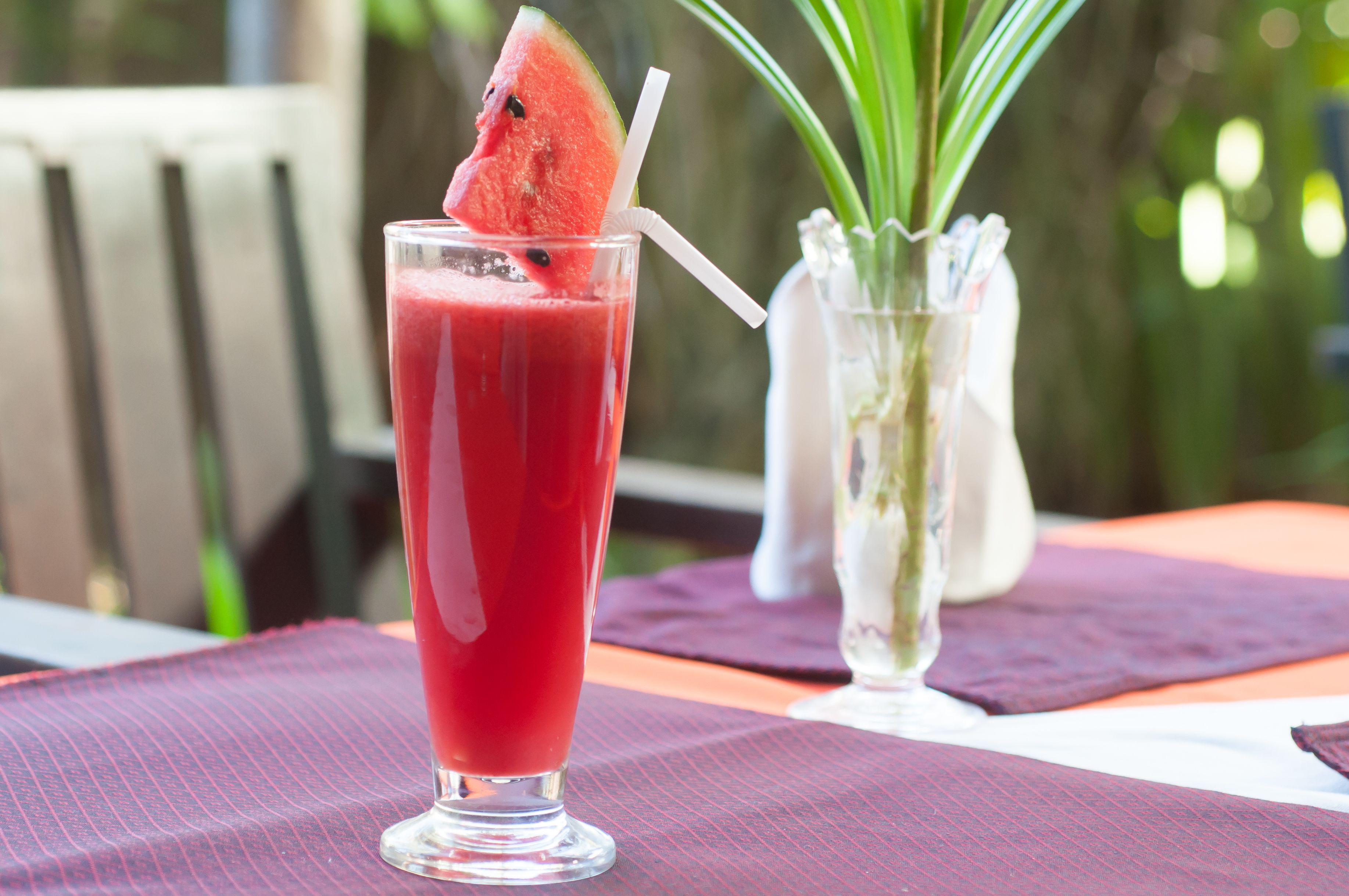 A tall glass of red beverage with a slice of watermelon on the rim, placed on a table with a purple placemat.