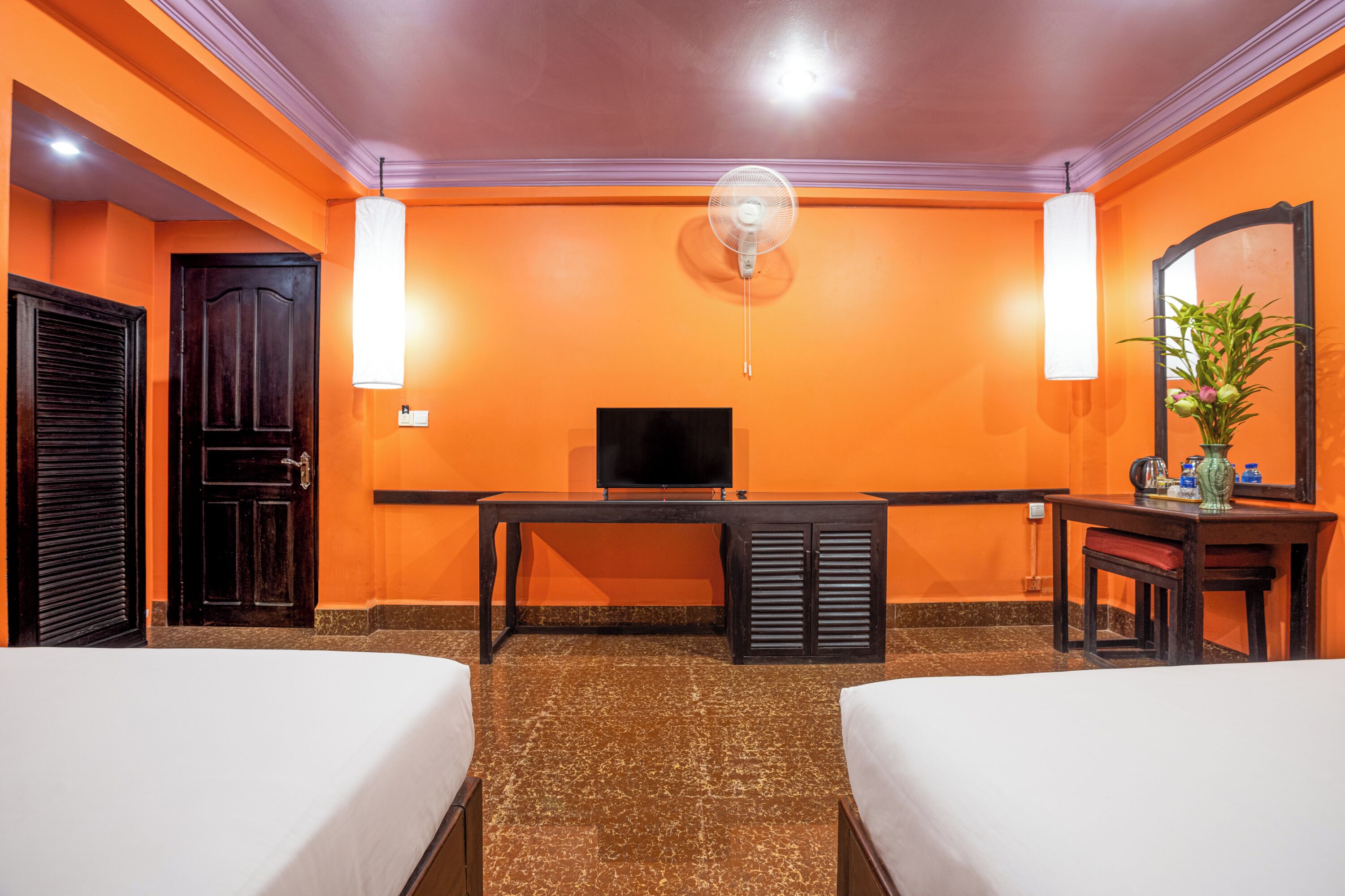 A hotel room with two beds, a desk, a television, a fan, and a mirror.