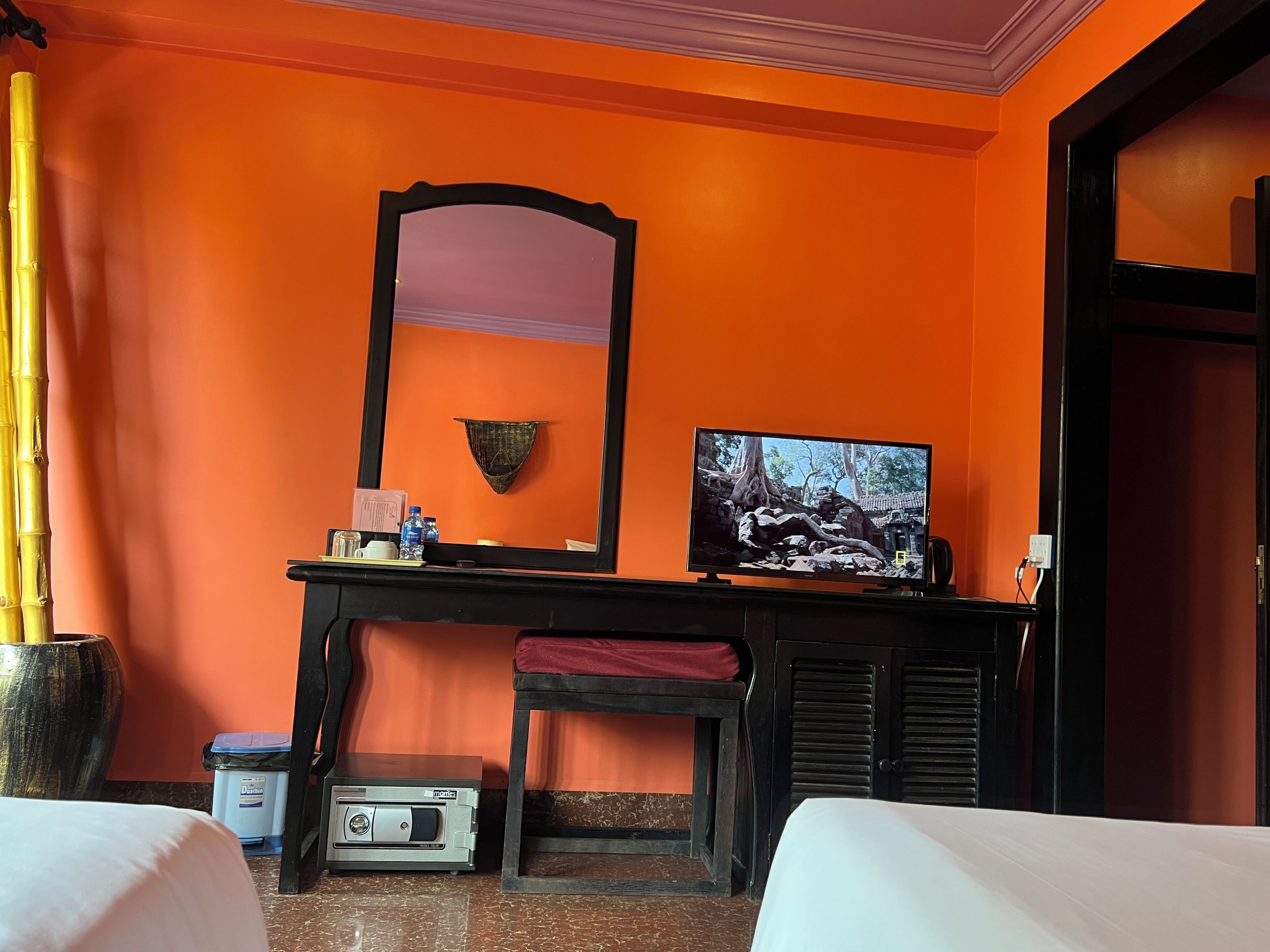 A hotel room with orange walls, a large mirror, a flat-screen TV, a desk with a red chair, and two beds.