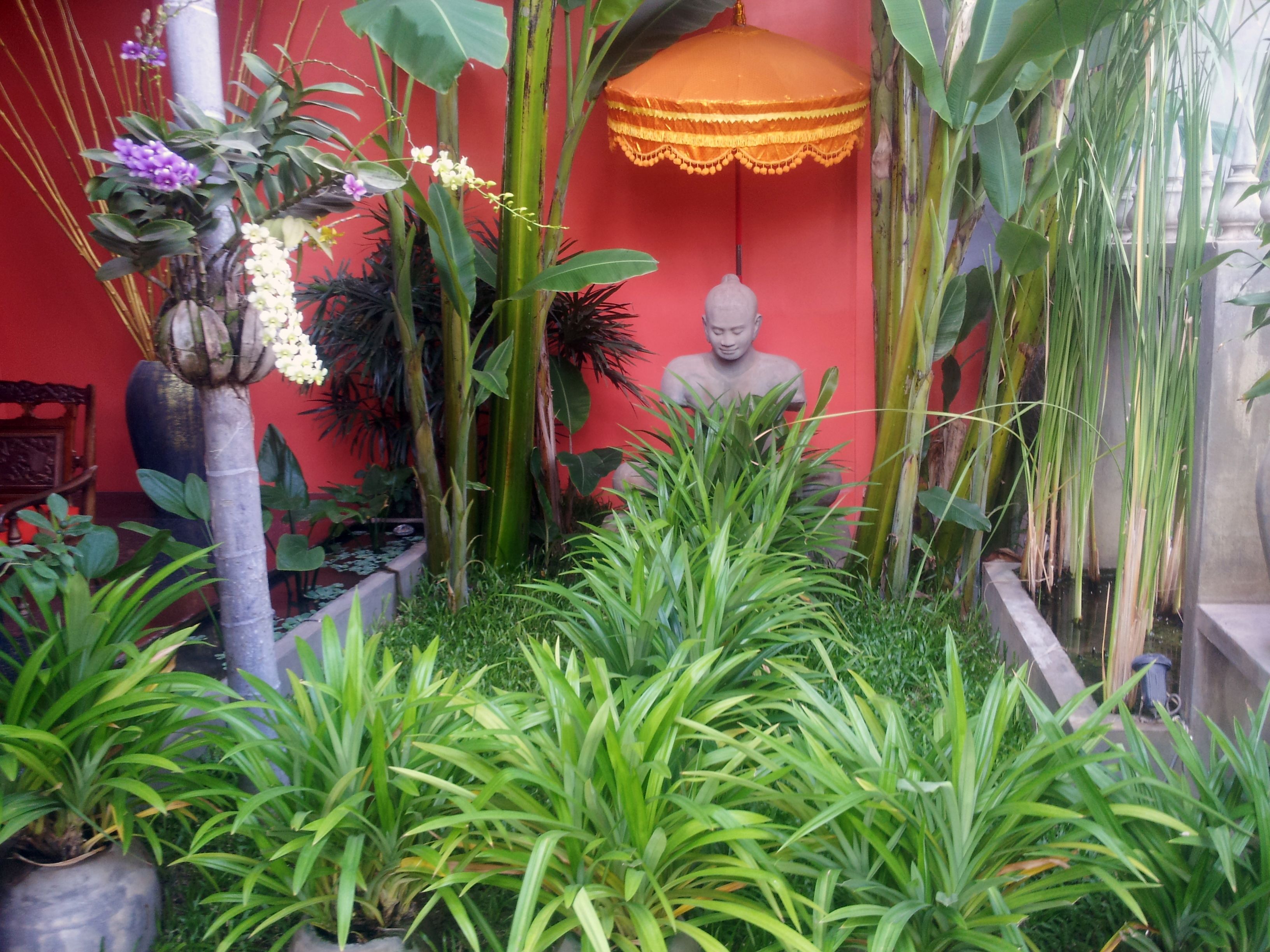 A serene indoor garden with a Buddha statue, lush greenery, and a decorative orange umbrella.
