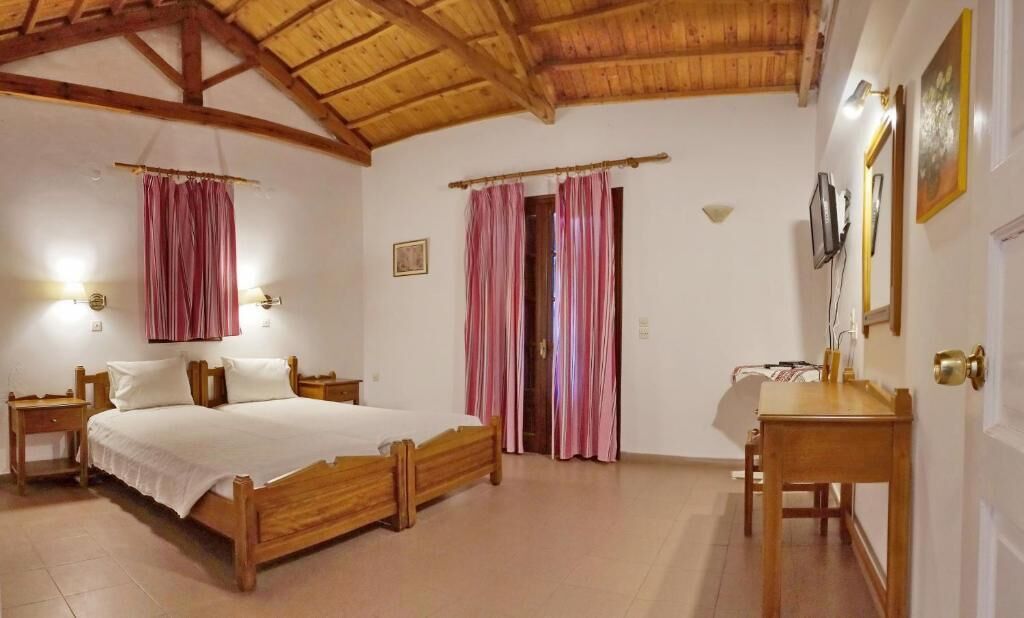 A bedroom with a wooden bed, two bedside tables, a desk, and a chair. There are two curtains and a door with a mirror and picture on the wall.
