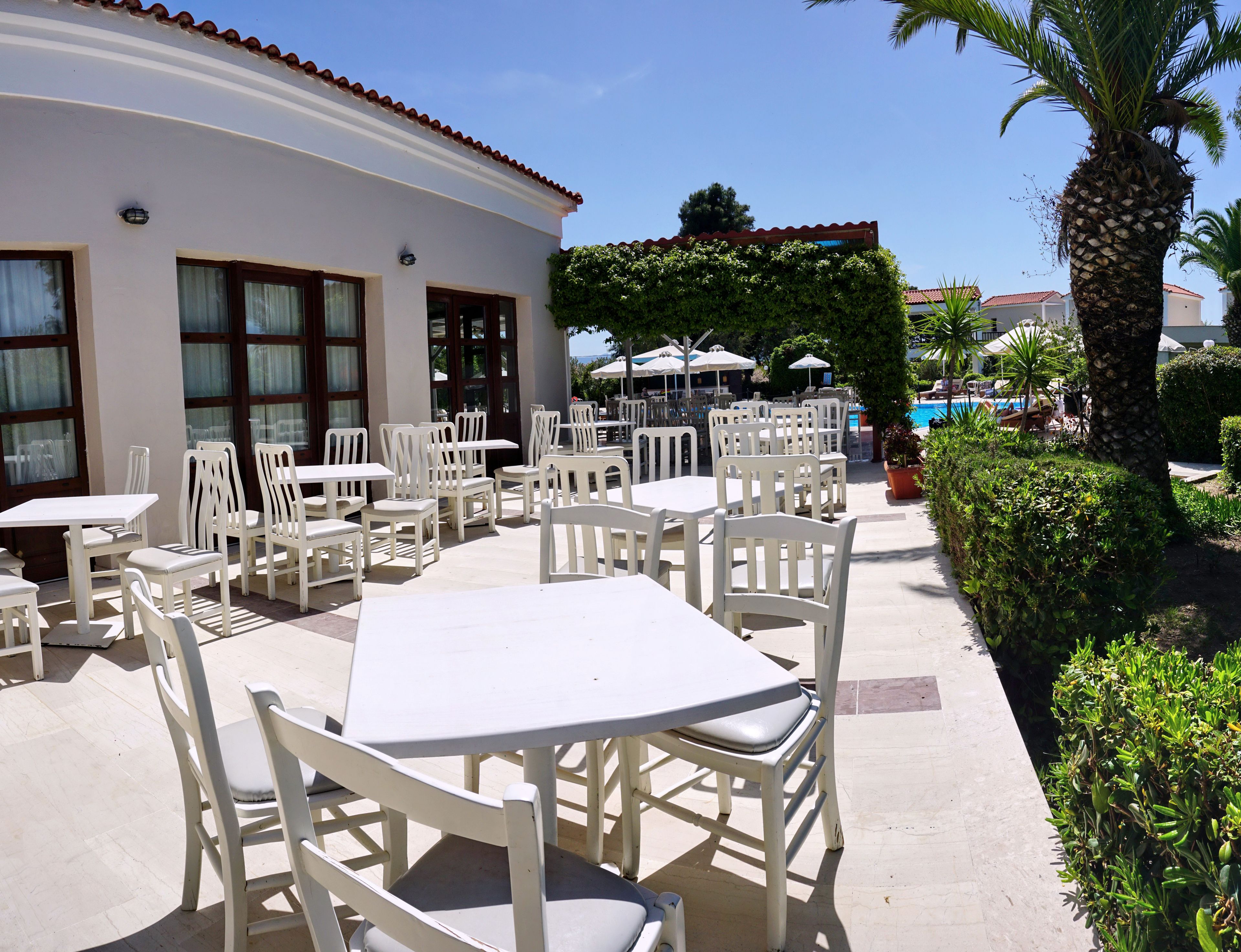 Un patio con mesas y sillas blancas, una piscina y un edificio con techo de tejas.