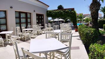 Un patio con mesas y sillas blancas, una piscina y un edificio con techo de tejas.