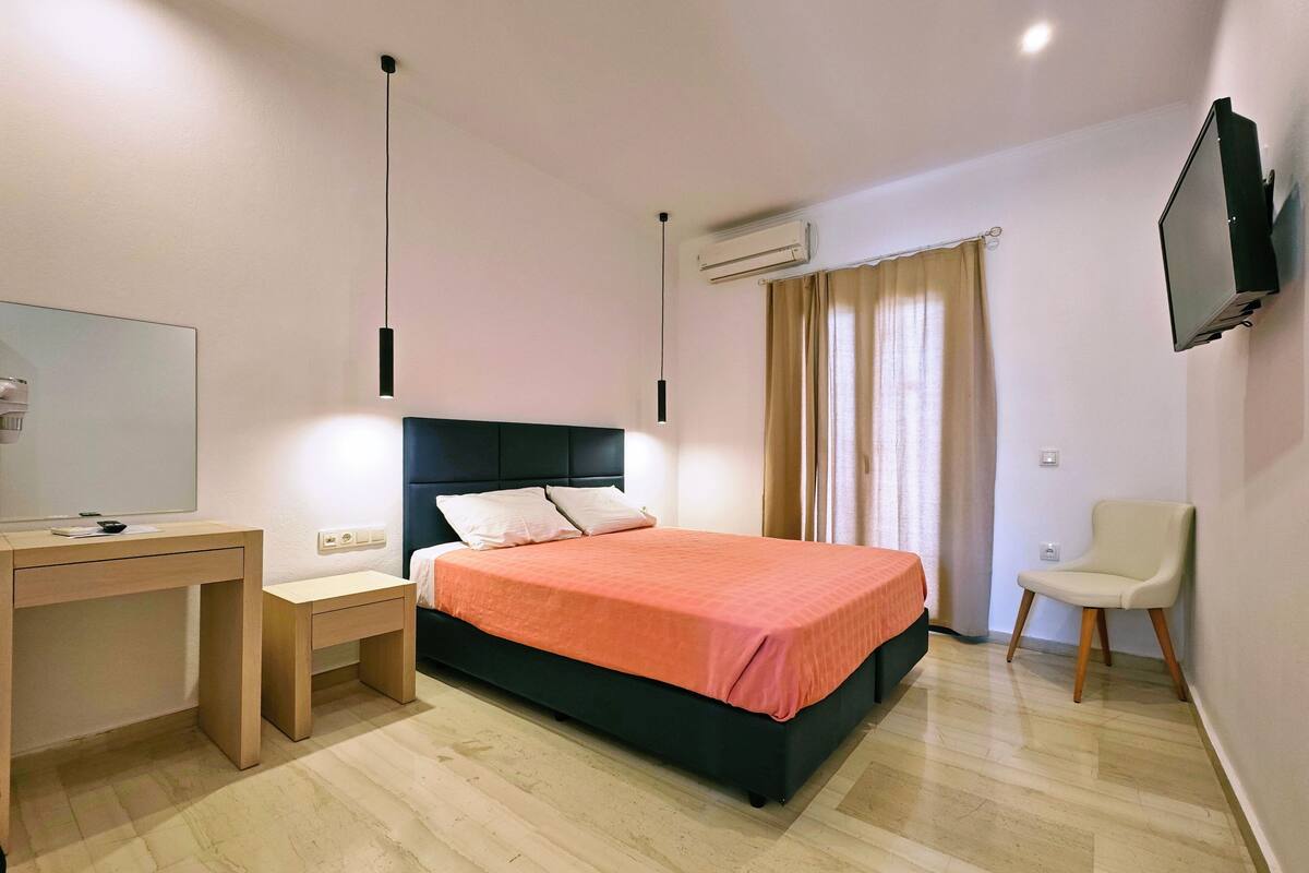A bedroom with a bed, a desk, a chair, a TV, and an air conditioning unit.