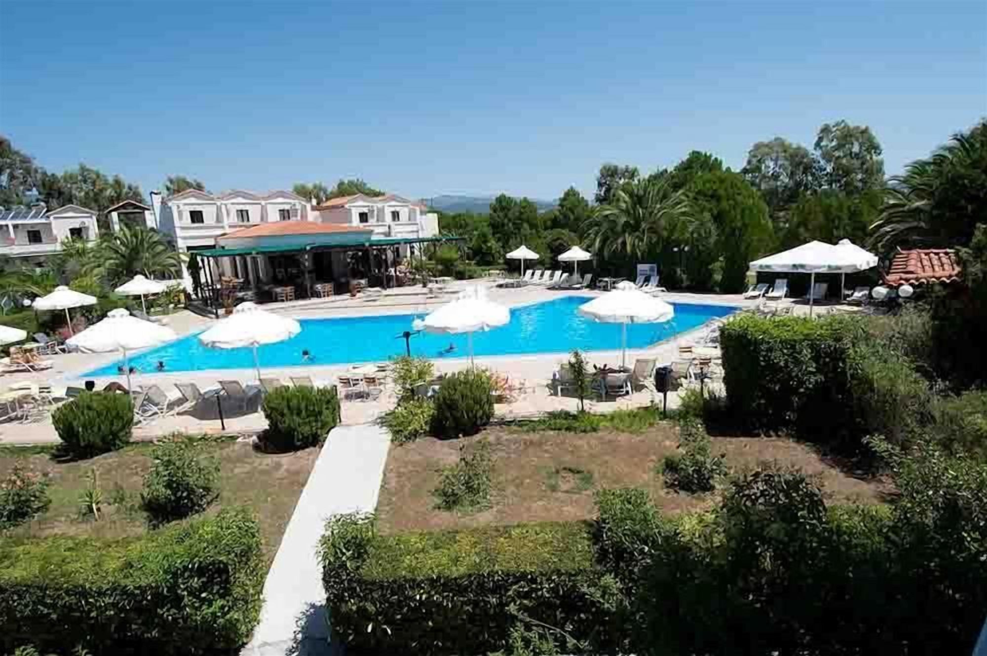 Zona de piscina con sillones de descanso y sombrillas, rodeada de vegetación y edificios.