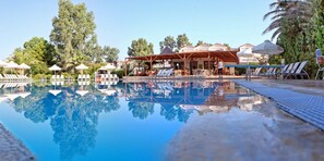 A pool with clear water reflecting trees, surrounded by lounge chairs and umbrellas. - Pasiphae (Lesvos)