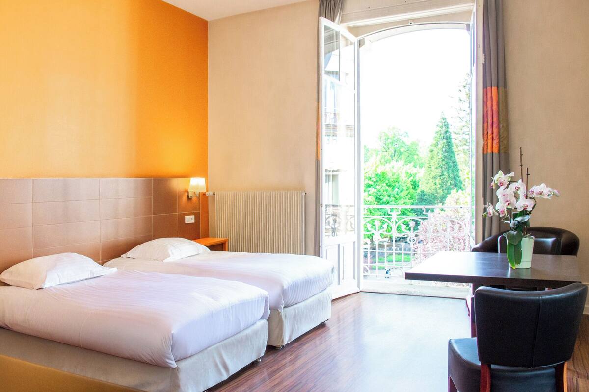 Habitación de hotel con dos camas, una mesa, una silla y un balcón con vistas a zonas verdes.