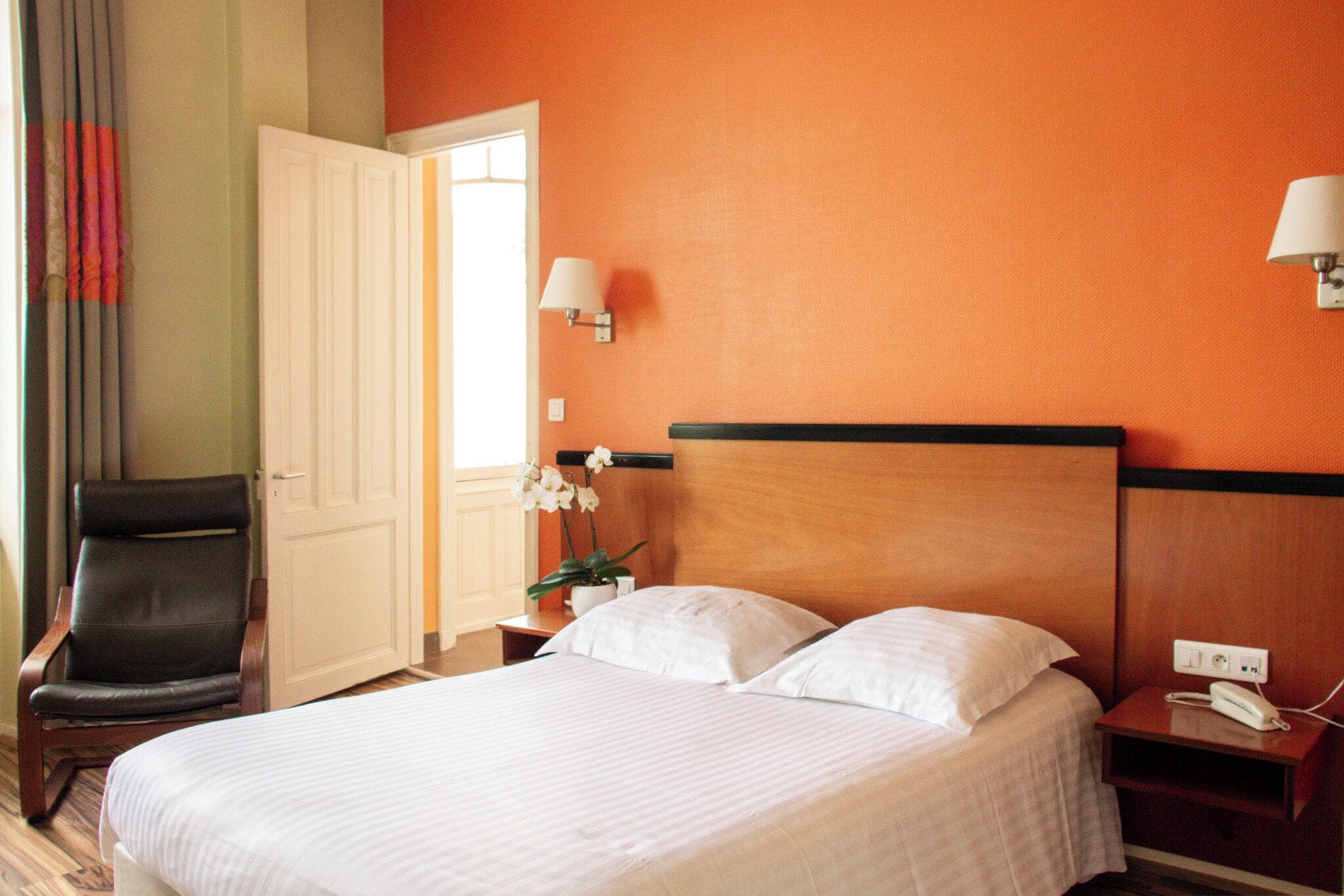 A hotel room with a bed, a chair, a nightstand, a lamp, and a door.