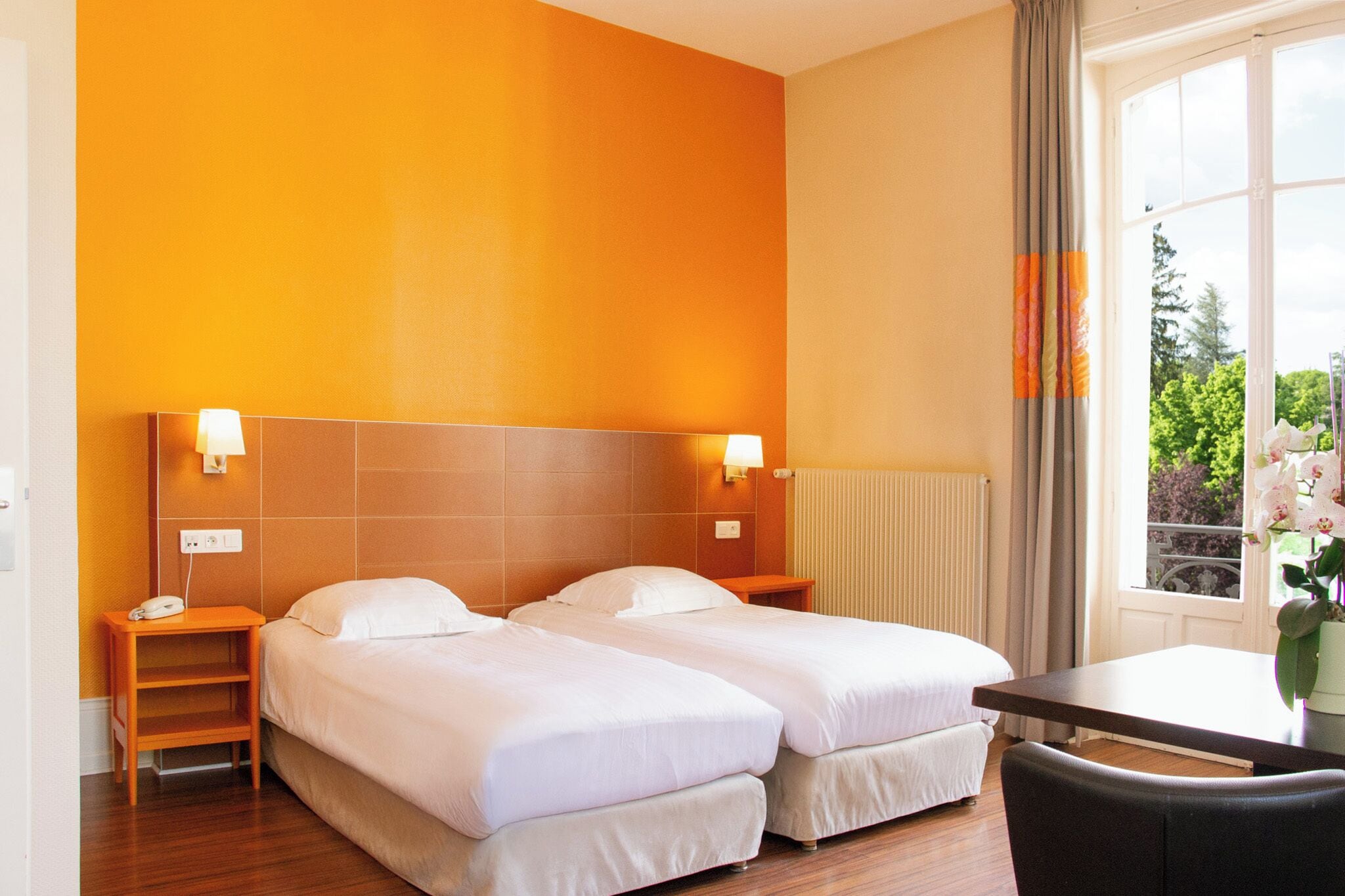 A hotel room with two beds, a wooden nightstand with a phone, a table, and a view of greenery through a window with curtains.