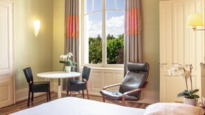 Hotelkamer met een bed, een eettafel met stoelen, een leren fauteuil, een raam met gordijnen en een potorkidee.