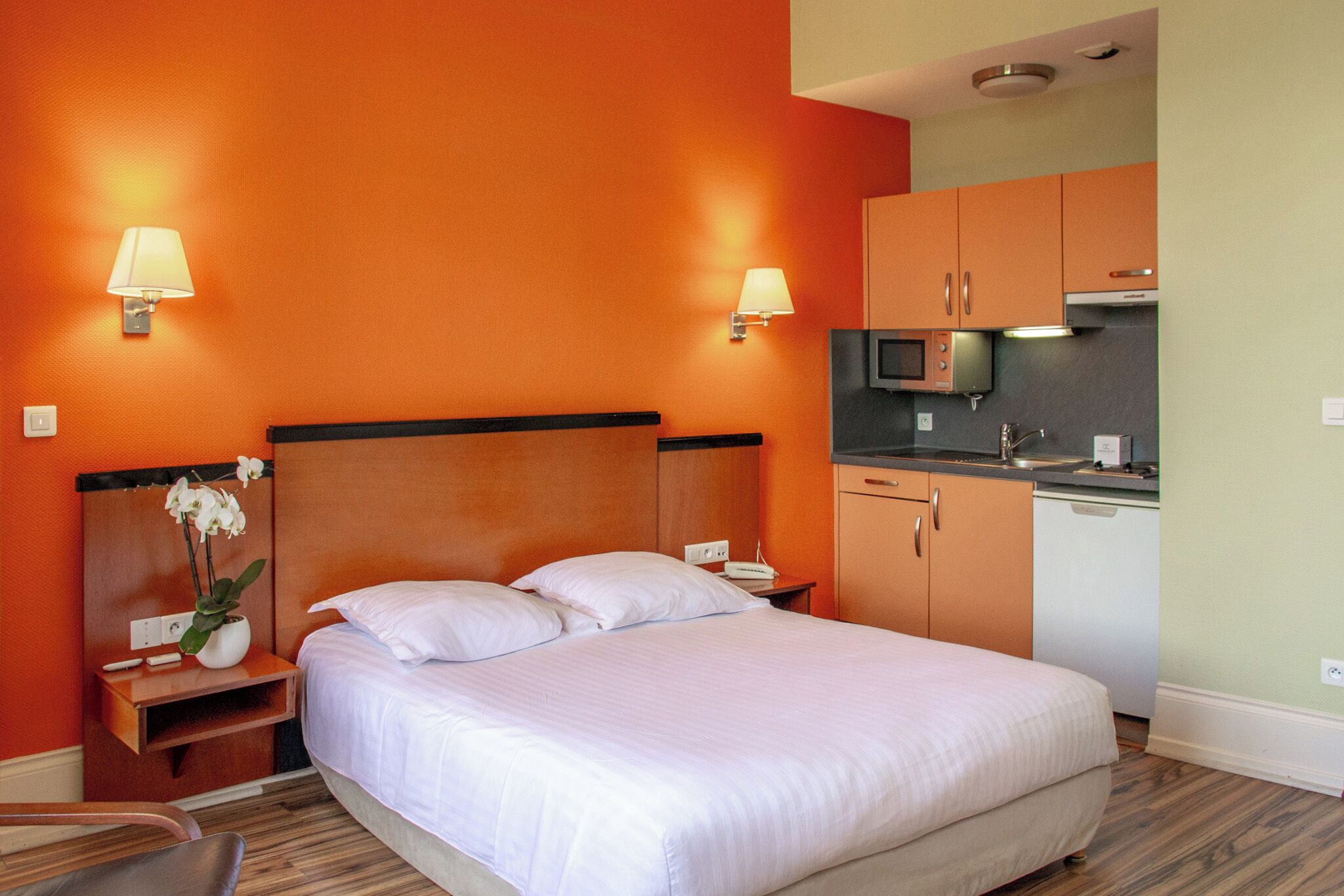 A hotel room with a large bed, a nightstand with a lamp, a small table with a vase of flowers, and a kitchenette with a microwave and sink.
