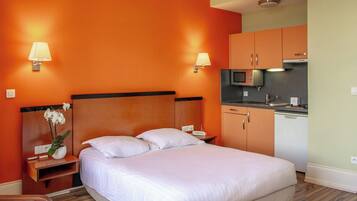 A hotel room with a large bed, a nightstand with a lamp, a small table with a vase of flowers, and a kitchenette with a microwave and sink.