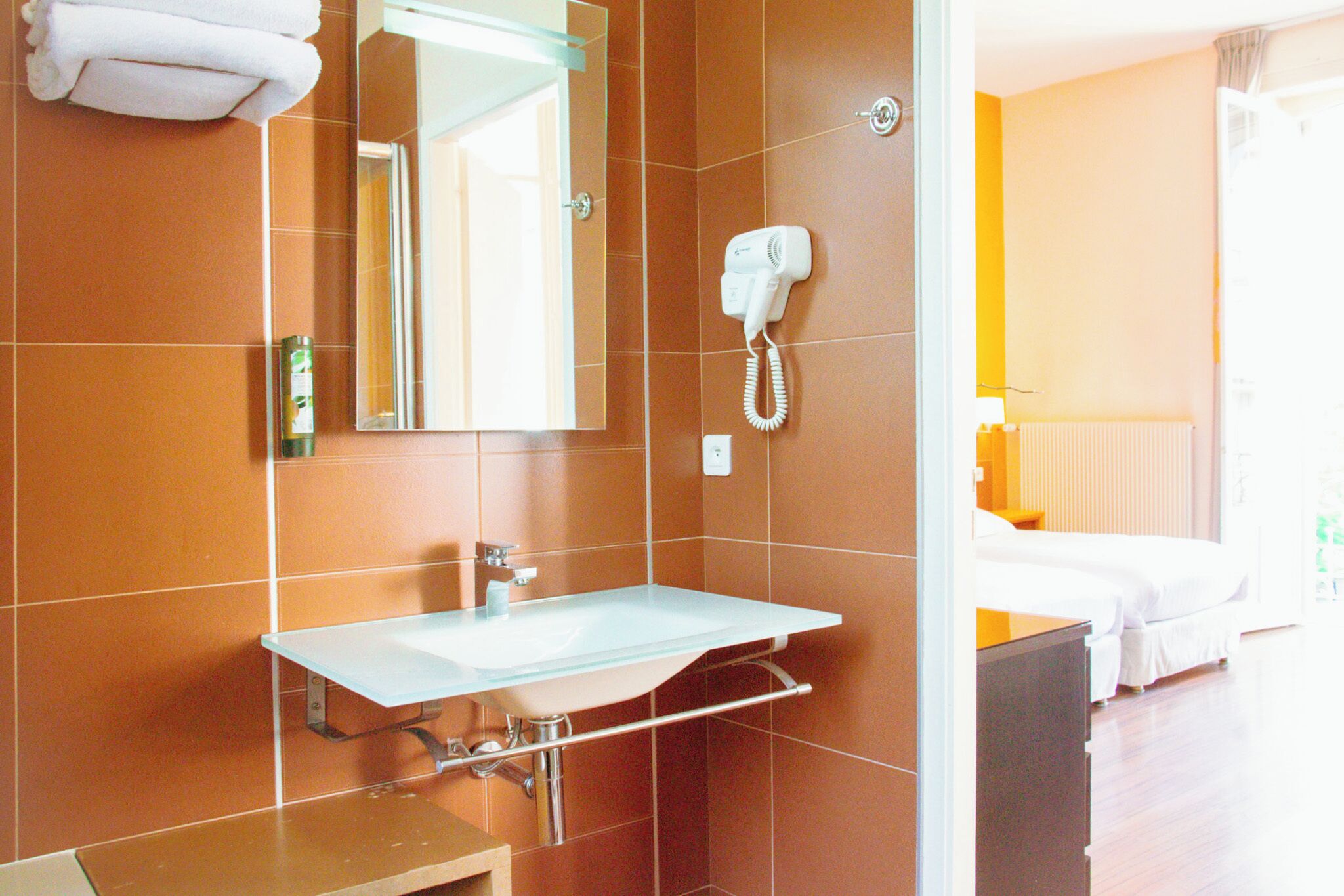 A hotel bathroom with a white sink, a hairdryer mounted on the wall, and a bed visible in the adjacent room.