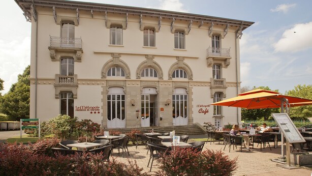 Een historisch gebouw met een caféterras, zitplaatsen buiten en een bord waarop staat dat het een toeristenverblijf is.