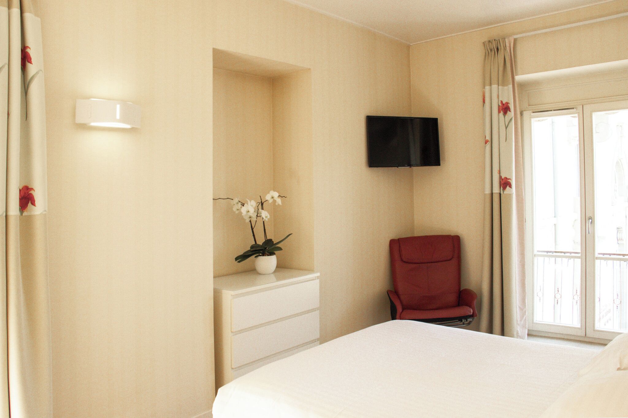 A hotel room with a bed, a red chair, a TV mounted on the wall, and a window with curtains.