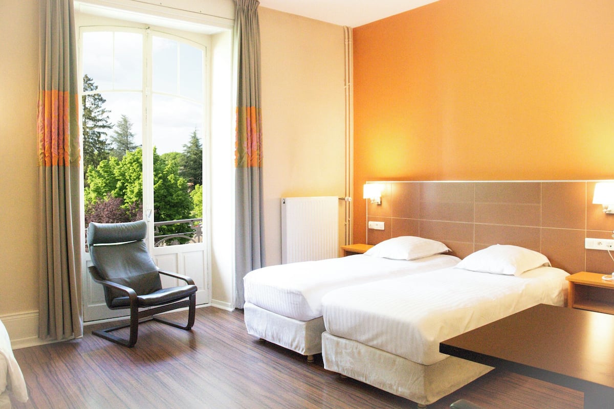 Habitación de hotel con dos camas, una silla, ventana con cortinas y vistas a zonas verdes.