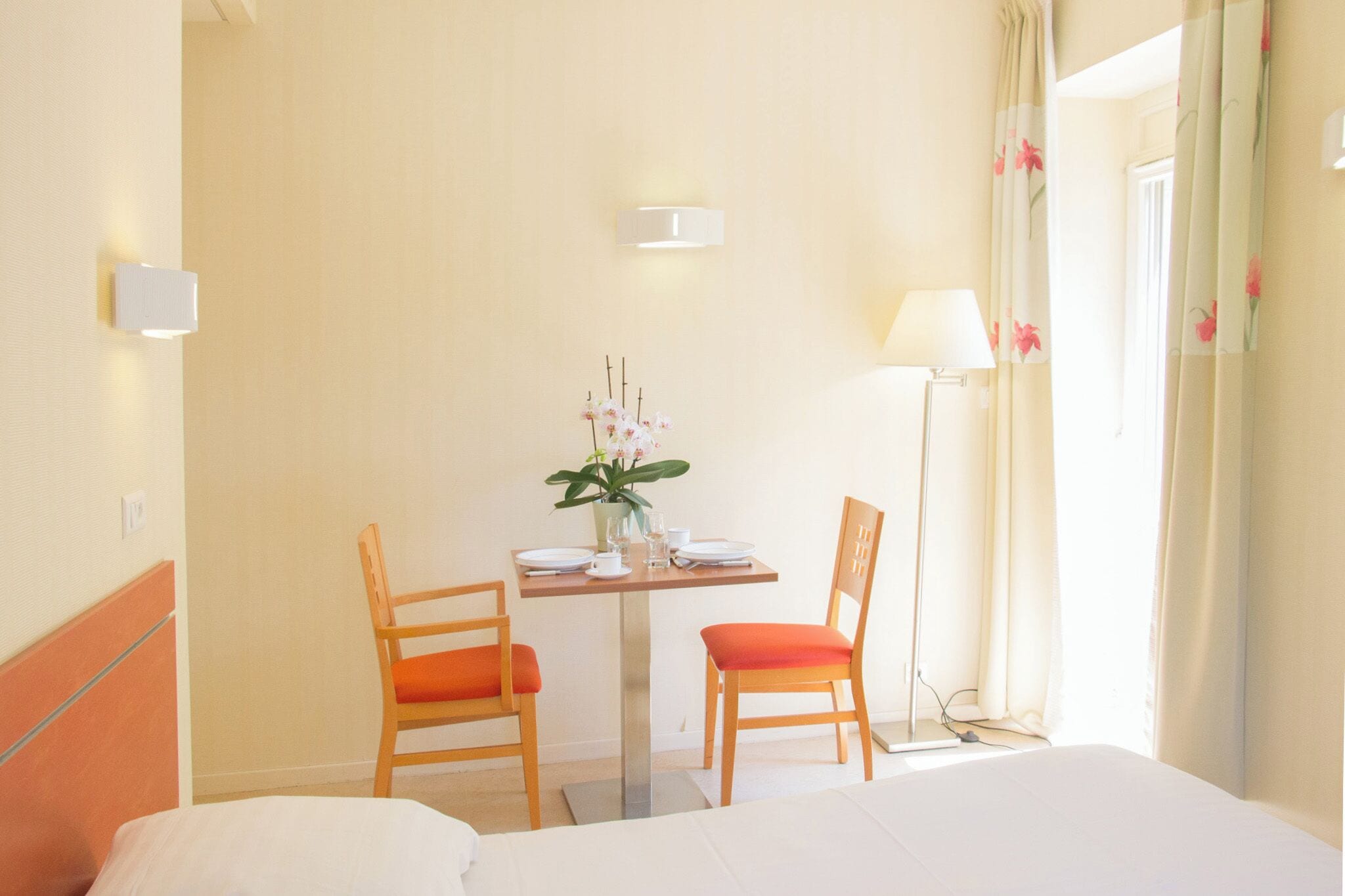 Una habitació d'hotel amb llit, una taula i cadires, un vas amb flors, una làmpada i una finestra amb cortines.