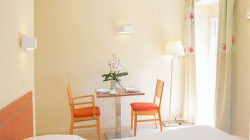 Una habitació d'hotel amb llit, una taula i cadires, un vas amb flors, una làmpada i una finestra amb cortines.