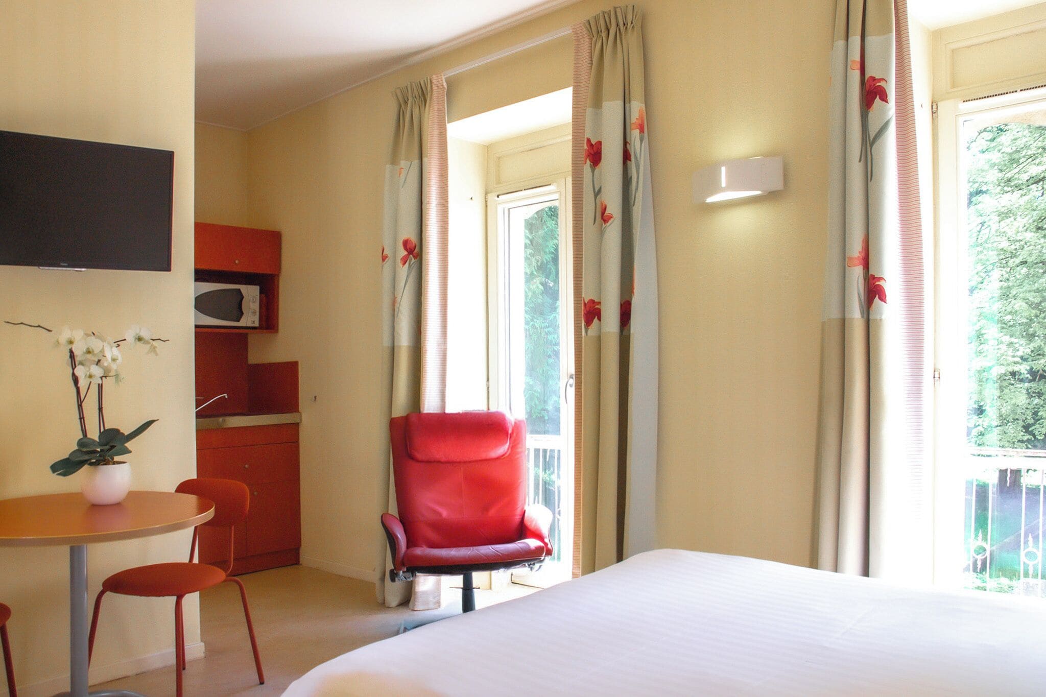 A hotel room with a bed, a red armchair, a small table with a plant, a microwave, and curtains with a floral pattern.