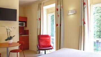 A hotel room with a bed, a red armchair, a small table with a plant, a microwave, and curtains with a floral pattern.