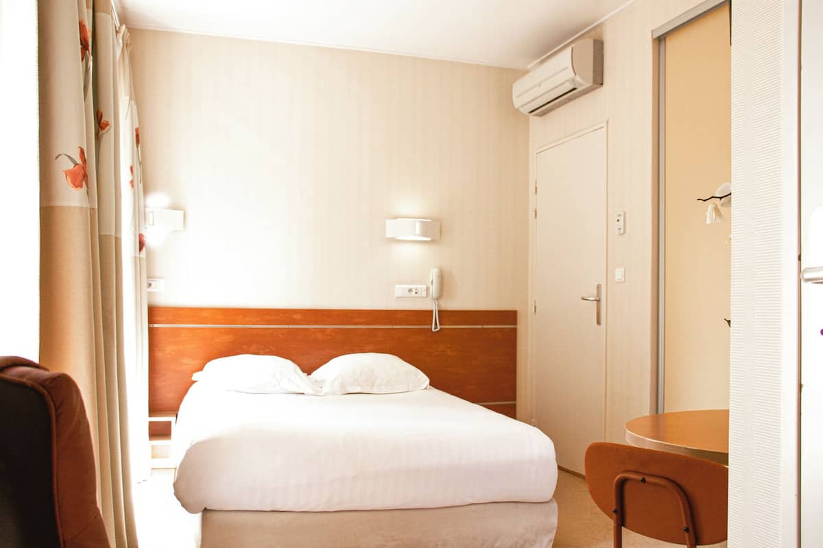 Una habitación de hotel con cama, una silla, una mesa, un teléfono en la pared y un equipo de aire acondicionado.