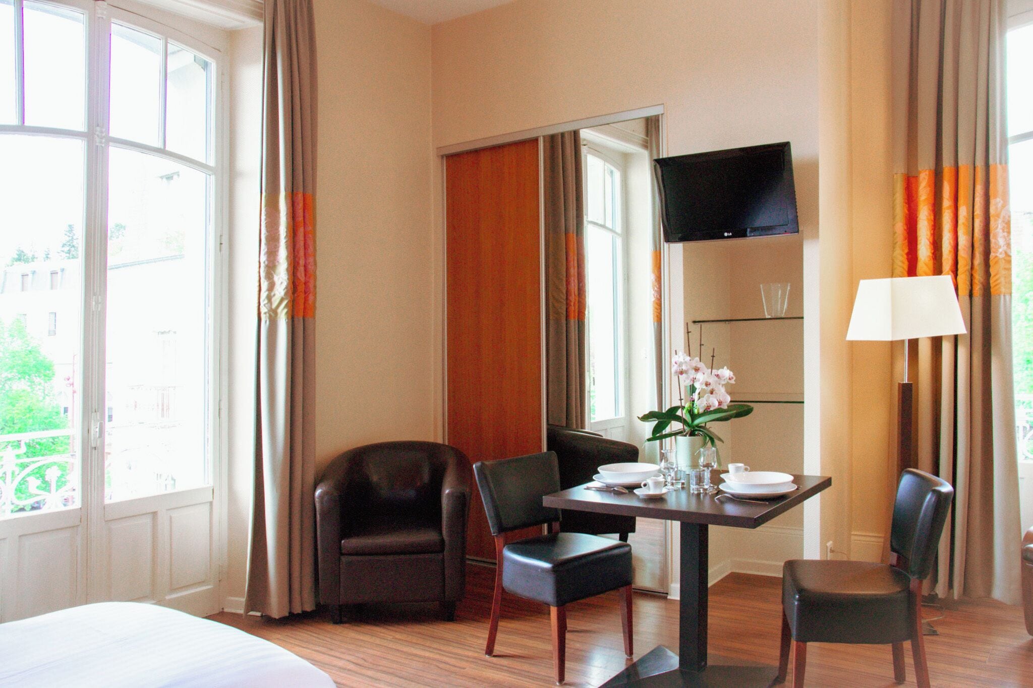 A hotel room with a bed, a dining table with chairs, a TV mounted on the wall, and a window with curtains.