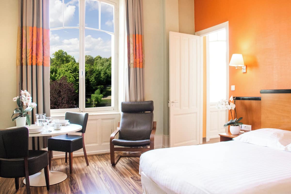 Habitación de hotel con cama, una mesa de comedor con sillas, un sillón, una ventana con cortinas y una mesita de noche con lámpara.