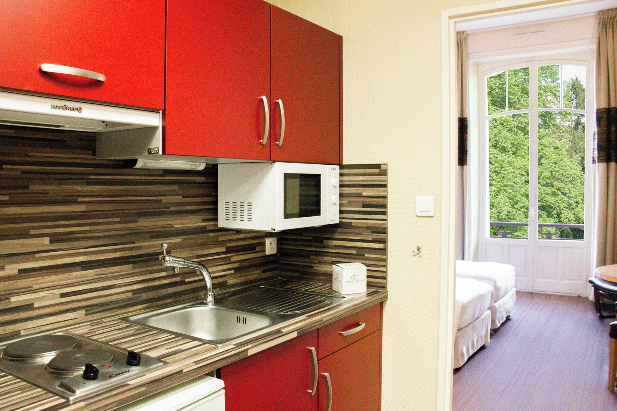 A compact kitchen with a microwave, sink, and stove, adjacent to a balcony with a view of greenery.