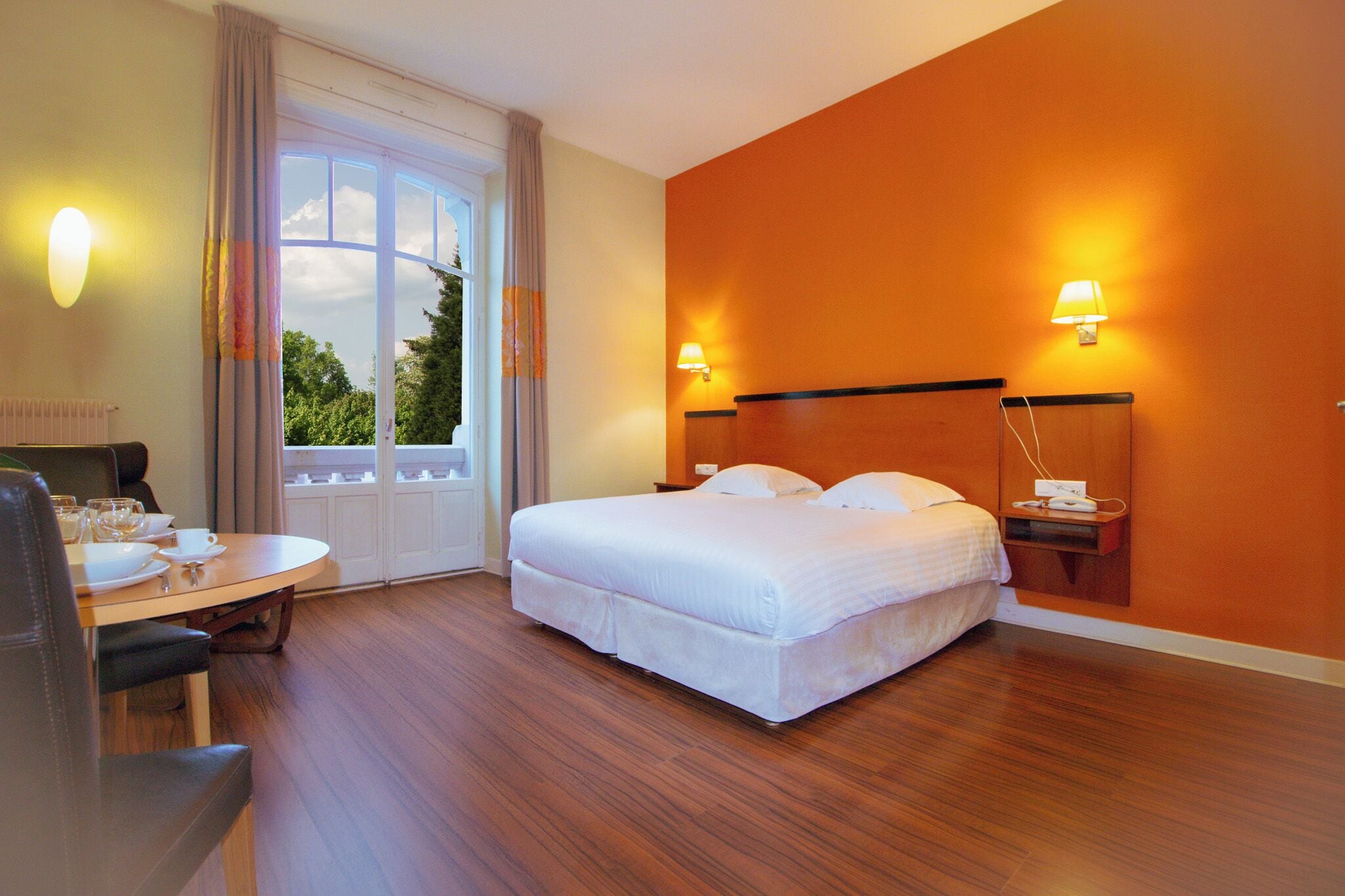 Une chambre d’hôtel comprenant un lit, un bureau avec une chaise, une table avec une chaise et une fenêtre avec des rideaux.