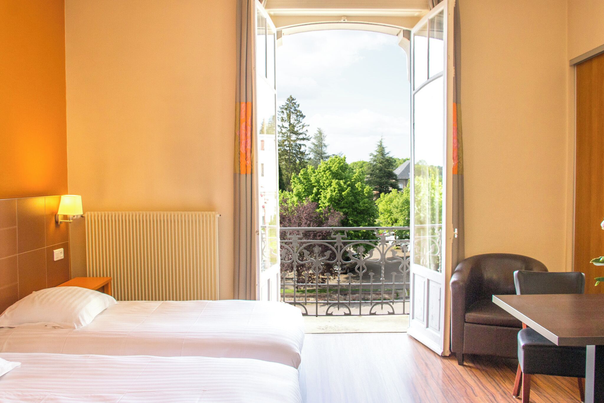 A hotel room with a bed, a table, a chair, and a balcony with a view of trees and a railing.