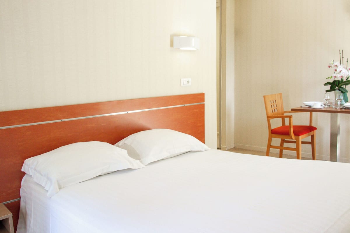 Habitación de hotel con una cama, una mesita de noche de madera, una silla y una pequeña mesa con un florero.