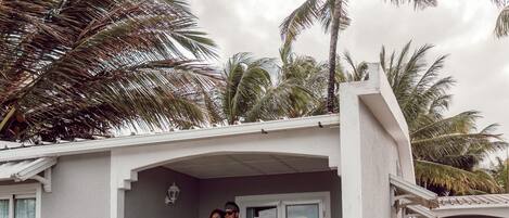 A two-story house with a white exterior, a porch with white railings, and a small garden area with tropical plants.