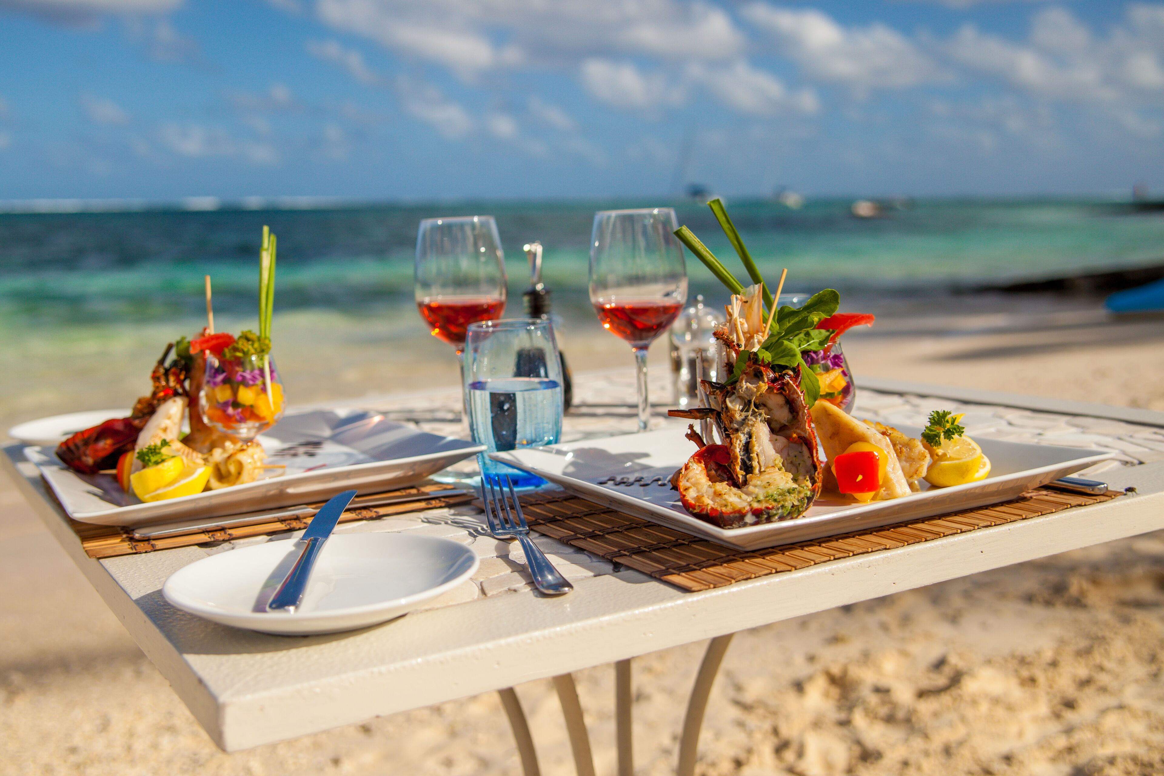 Una mesa servida con platos de comida, copas de vino tinto y una botella de agua en una playa con el océano al fondo.