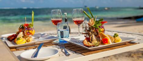 Ein Tisch ist gedeckt mit Speisen, Weingläsern und einer Flasche Wasser, mit Blick auf den Strand und den Ozean.